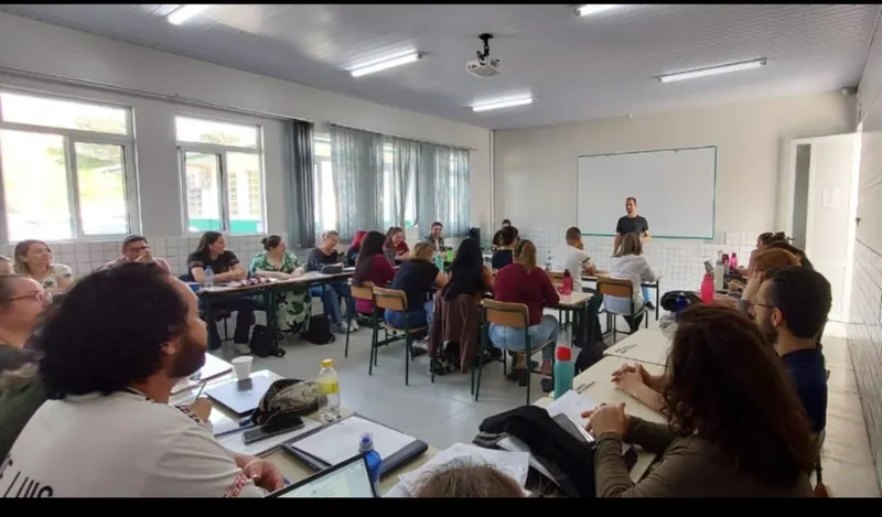 Sala de aula cheia de alunos e professores sentados, com Edilson ao fundo, de pé, durante uma palestra