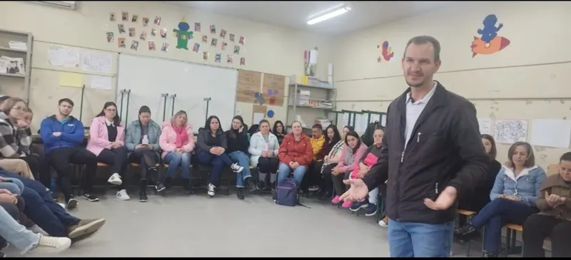 Edilson de pé, falando com os braços abertos, diante de uma roda de pessoas sentadas numa sala de escola