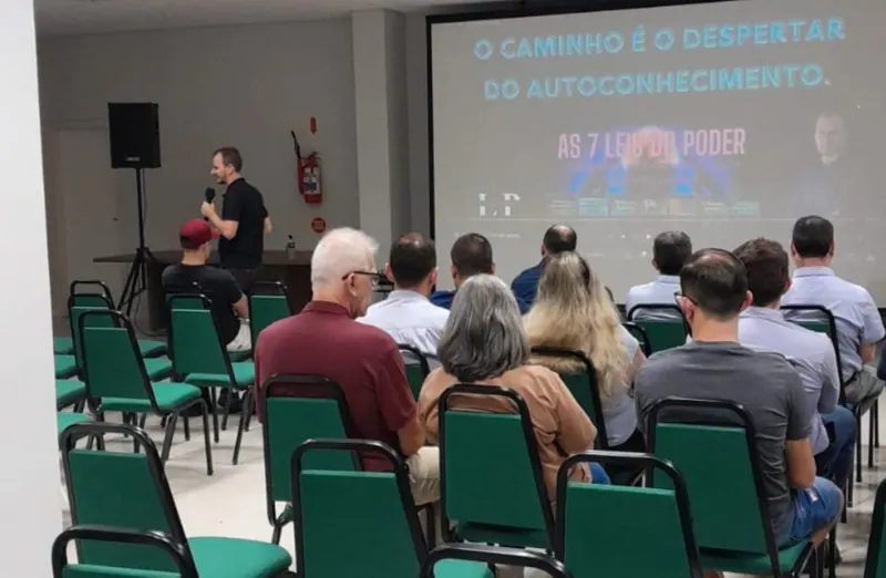 Edilson com o microfone na mão, diante do público sentado e de um telão com a frase O caminho é o despertar do autoconhecimento